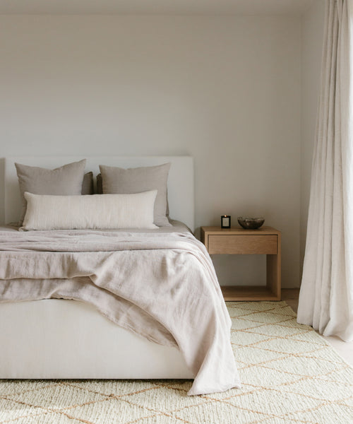 Minimalist bedroom with a neatly made bed in neutral tones, beige bedding, and pillows. A wooden nightstand with a bowl and candle sits beside the bed. Soft light filters through curtains onto a Wilder Diamond Jute Rug.