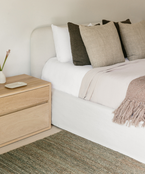 A neatly made Cove Bed with white and beige Belgian Libeco linen bedding, neutral throw pillows, and a tan throw blanket sits beside a light wood nightstand holding a small white vase and plate, all atop a textured green rug.