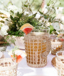 The Seagrass Picnic Pitcher, featuring a woven wicker cover and filled with a light brown drink topped with grapefruit and herbs, sits alongside matching seagrass glassware and green floral decor on a sunlit woven tabletop.
