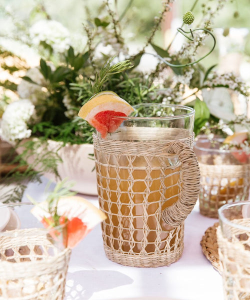 The Seagrass Picnic Pitcher, featuring a woven wicker cover and filled with a light brown drink topped with grapefruit and herbs, sits alongside matching seagrass glassware and green floral decor on a sunlit woven tabletop.