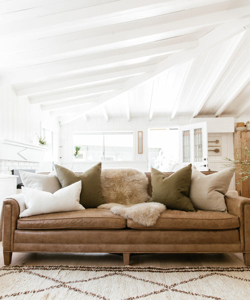 A brown leather sofa with neutral and olive green pillows, including the Linen Lumbar Pillow and a soft sheepskin throw, sits in a bright white living room with vaulted ceilings, patterned rug, and ample natural light.