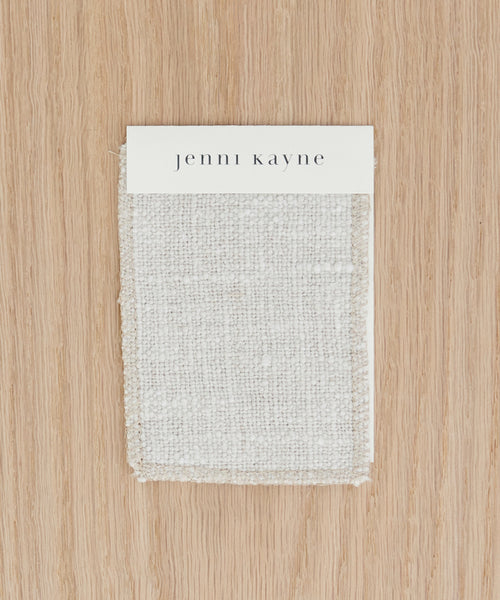 A rectangular Fabric Swatch Textured Natural Linen, made from Belgian flax linen, is displayed on a light wood surface with a card labeled jenni kayne in lowercase black letters attached at the top.