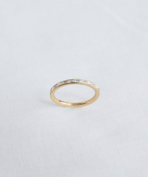 The June Baguette Band, crafted in 14k gold with a row of clear rectangular stones, evokes the style of a diamond baguette band and is shown on a white textured background.
