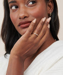 A woman with medium-dark skin and shoulder-length hair rests her hand on her cheek, displaying the June Baguette Band in 14k gold set with small gemstones. She wears a white top and a small hoop earring, embodying Jenni Kayne’s classic jewelry style.