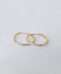 A pair of June Baguette Hoops in 14k gold with small rectangular clear stones set along the front, shown on a light textured surface.