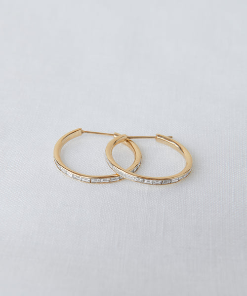 A pair of June Baguette Hoops in 14k gold with small rectangular clear stones set along the front, shown on a light textured surface.