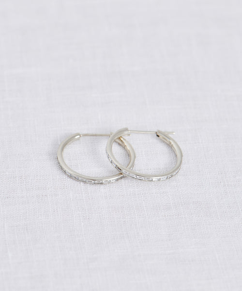 A pair of June Baguette Hoops in white gold, featuring a simple, elegant design and smooth, shiny finish, displayed on a light textured fabric background. Inspired by the timeless style of Jenni Kayne jewelry.