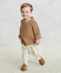 A young child with light brown hair smiles while standing indoors, wearing the cozy Kids Fisherman Sweater, off-white pants, and brown shoes on a light carpet with a neutral background.