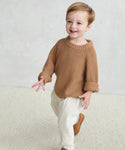A young child with light brown hair smiles while walking indoors on a light carpet, dressed in a cozy Kids Fisherman Sweater, white pants, and tan shoes.