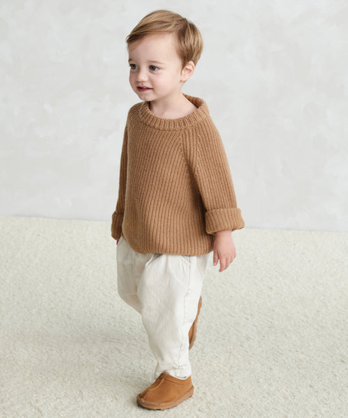 A young child with short light brown hair wears a Kids Fisherman Sweater, cream pants, and brown shoes, standing on a light textured floor against a neutral background.