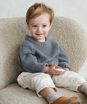 A smiling young child with light brown hair wears a gray Kids Fisherman Sweater, cream pants, and brown shoes while sitting on a textured cream chair and holding an object.
