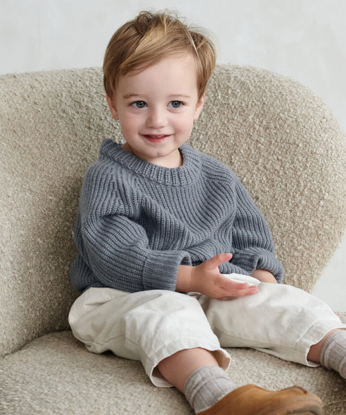 A smiling young child with light brown hair wears a gray Kids Fisherman Sweater, cream pants, and brown shoes while sitting on a textured cream chair and holding an object.