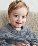 A young child with light brown hair and blue eyes smiles while wearing the Kids Fisherman Sweater, sitting on a soft, light-colored chair.