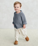 A smiling young child with short light brown hair wears the Kids Fisherman Sweater in dusty blue, paired with cream pants and brown shoes, standing on a light carpet against a pale textured background.
