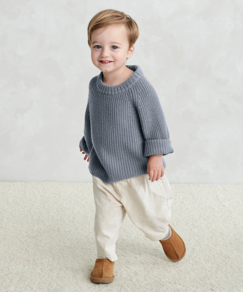 A smiling young child with short light brown hair wears the Kids Fisherman Sweater in dusty blue, paired with cream pants and brown shoes, standing on a light carpet against a pale textured background.