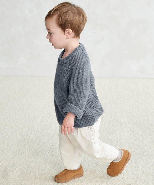 A young child with short light brown hair walks sideways on a light carpet, wearing the Kids Fisherman Sweater in gray, paired with cream-colored pants and brown slip-on shoes against a softly textured plain background.