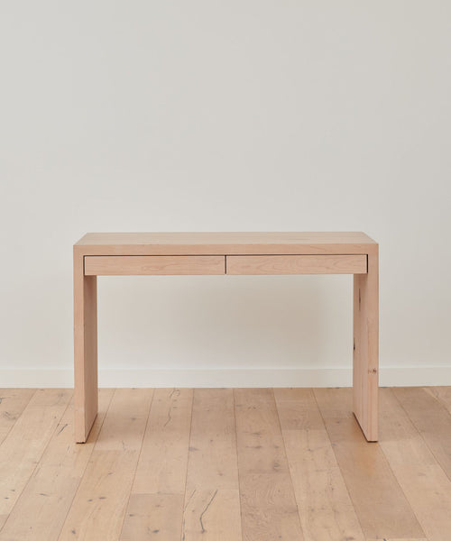 The Newport Desk is a minimalist solid wood piece with clean lines and two drawers, shown against a plain white wall on a matching wooden floor.