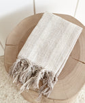 A folded Laurel Hand Towel—light beige, handwoven in Turkey from a linen cotton blend with fringed edges—rests on a round, natural wood stool. The background features a light-colored wall and floor.