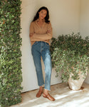 A woman stands indoors by a leafy plant wall and large potted plant, wearing a beige sweater, blue jeans, and the Leather Lake Sandal. Her arms are relaxed as she crosses one leg over the other, with sunlight streaming into the space.