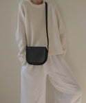 A person, head partially out of frame, poses in a cream knit sweater and loose white pants with the Small Leather Saddle Bag—handmade in Italy from black leather—set against a neutral background.