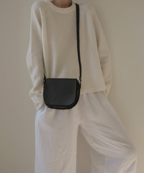 A person, head partially out of frame, poses in a cream knit sweater and loose white pants with the Small Leather Saddle Bag—handmade in Italy from black leather—set against a neutral background.