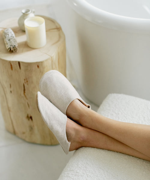 Someone wearing the Linen House Slipper rests their feet on a soft surface by a wooden stool holding a candle, sage bundle, and small vase, next to a white bathtub.