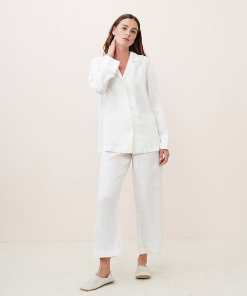 A woman with long straight hair poses naturally against a plain background, wearing a loose white button-up shirt, matching wide-leg pants, and Linen House Slippers.