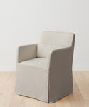 The Sunset Dining Arm Chair Slipcover in light beige linen, with a matching cushion and skirted design, covers an armchair on a light wooden floor against a plain white wall.