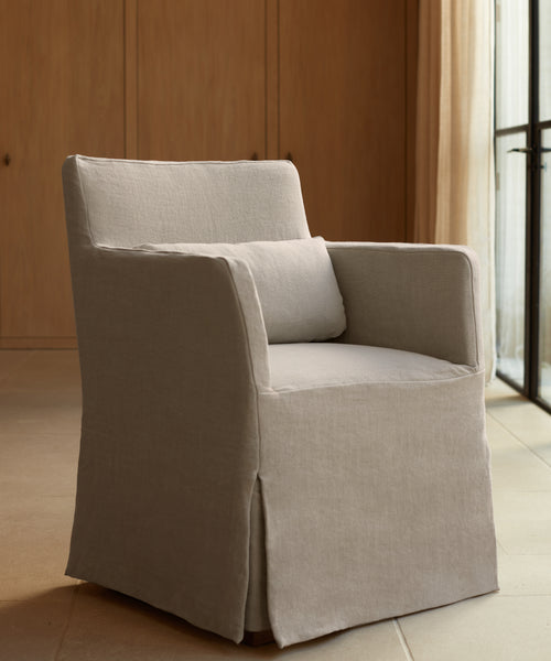 The Sunset Dining Arm Chair, featuring light beige fabric and a matching lumbar pillow, adds elegance to a neutral-toned room with wooden paneling and large windows.