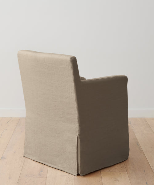 The Sunset Dining Arm Chair, featuring a beige fabric slipcover, is displayed from the back on a light wooden floor with a plain white wall backdrop.