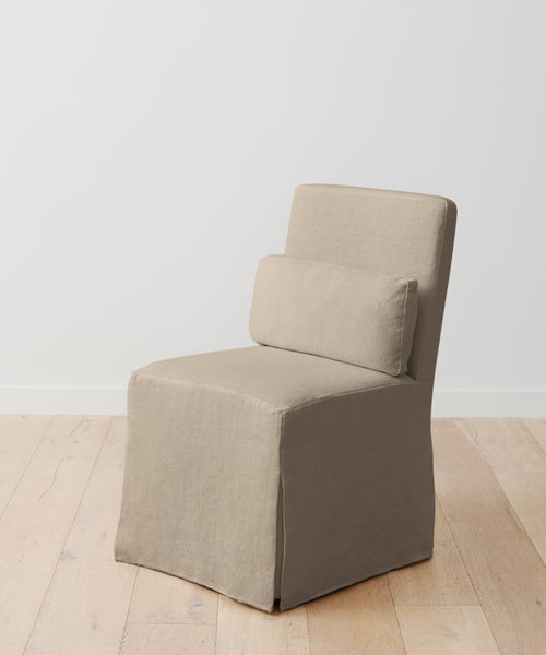 The Sunset Dining Chair Slipcover in taupe linen covers a beige armless chair with a rectangular backrest and cushion, shown on a light wooden floor against a plain white wall.