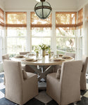 A bright dining room features a round table set for six with beige chairs accented by Sunset Dining Chair Slipcovers. Large windows with bamboo shades fill the space with light, while a vase of white flowers sits at the center of the table.