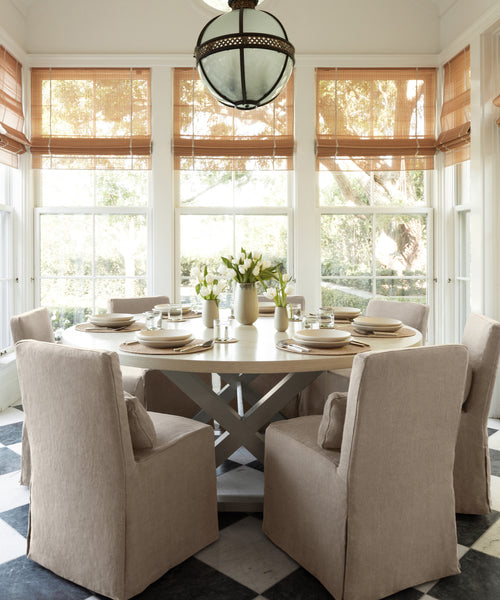 A bright dining room features a round table set for six with beige chairs accented by Sunset Dining Chair Slipcovers. Large windows with bamboo shades fill the space with light, while a vase of white flowers sits at the center of the table.