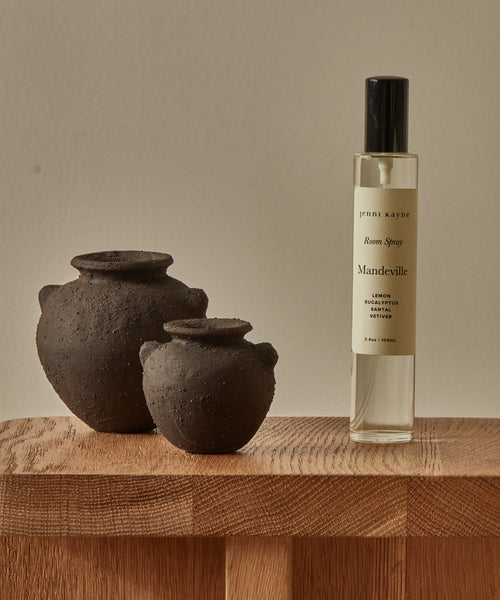 A clear bottle of Mandeville Room Spray stands on a wooden surface next to two small round black clay pots, set against a neutral background.