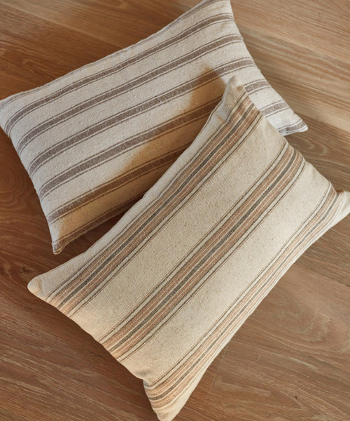 Two Mill Pillows—rectangular, beige with horizontal brown and cream stripes, feather down inserts—overlap slightly as they rest on a wooden floor, revealing the wood grain beneath.