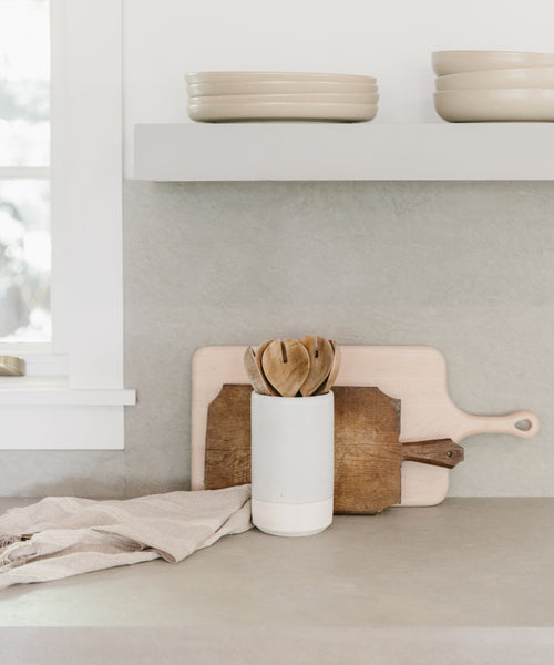 A minimalist kitchen counter with a Thin Ceramic Vessel holding wooden utensils, two cutting boards against the backsplash, stacked handmade ceramics on a shelf, and a folded beige cloth beside the vase.