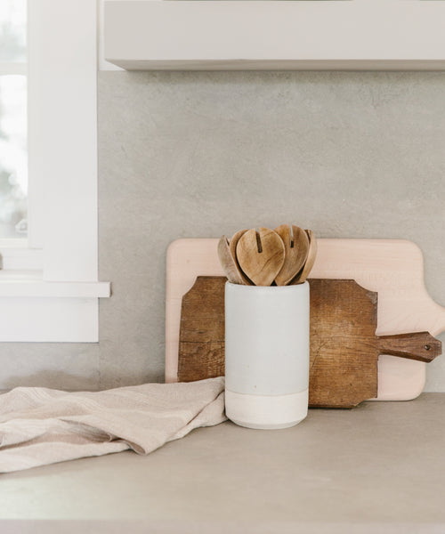 A neutral-toned kitchen countertop features a Thin Ceramic Vessel holding wooden spoons, two wooden cutting boards propped behind it, and a beige dish towel draped on the counter.