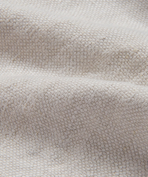 Close-up of the Cove Bed Slipcover, featuring light beige Belgian Libeco linen with a textured, soft surface and visible weave. The fabric is gently draped, forming subtle folds and shadows.