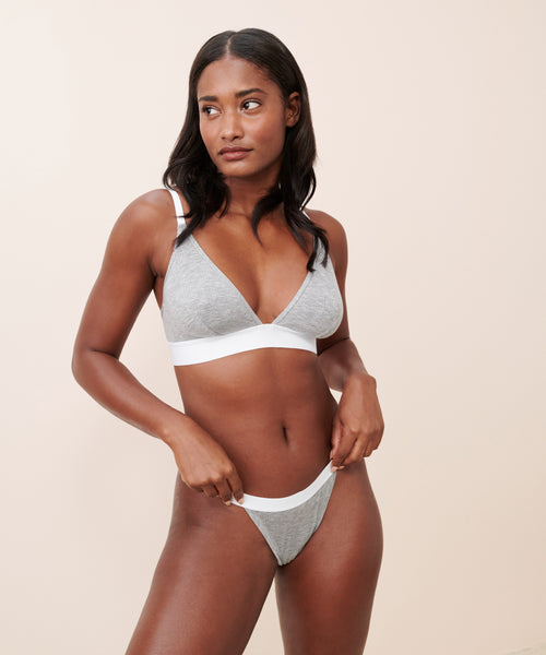 A woman stands against a plain beige background, looking to the side with her hand on her hip, wearing the Whipped Thong in grey micro modal with white trim.