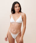 A woman stands against a neutral background wearing a white bralette and matching Whipped Thong, both with beige bands, looking to the side with a relaxed expression.