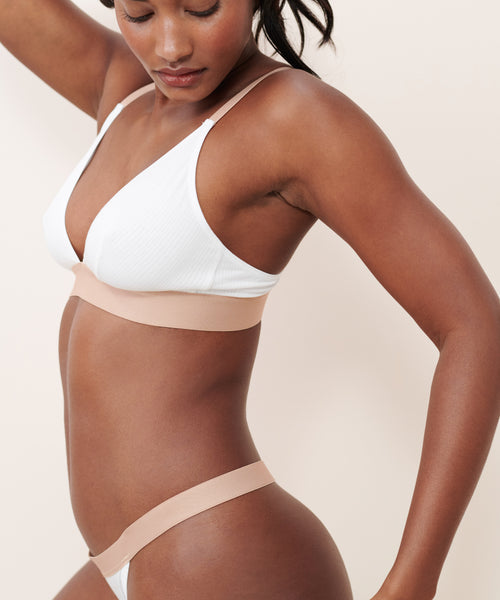 A woman in a white bralette and the Whipped Thong poses against a light background, one arm raised and head tilted down, showcasing the effortless style and comfort of this underwear.