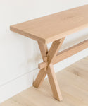The Oak Entry Table features a minimalist design with a light wood finish and crossed legs, inspired by Jenni Kayne furniture, and looks clean and modern against a white wall on a light wood floor.