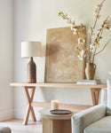 A light rustic Oak Entry Table features a ceramic lamp, framed abstract art, stacked books, a branch-filled vase, and a lit candle on a nearby round side table beside a pale sofa—reflecting Jenni Kayne’s signature style.