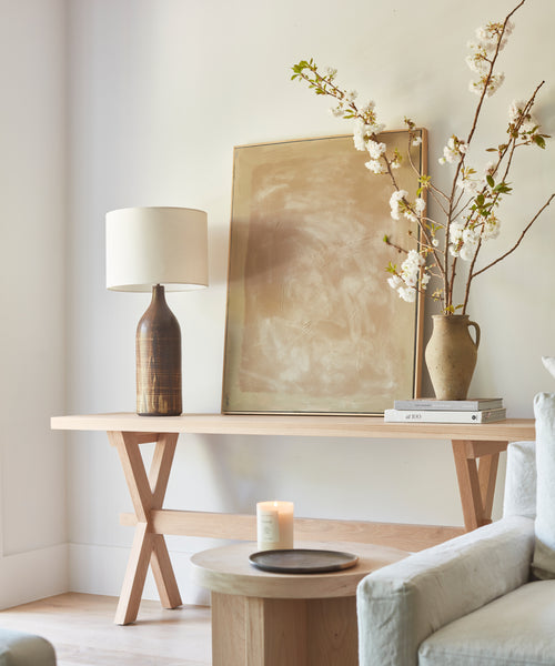 A light rustic Oak Entry Table features a ceramic lamp, framed abstract art, stacked books, a branch-filled vase, and a lit candle on a nearby round side table beside a pale sofa—reflecting Jenni Kayne’s signature style.