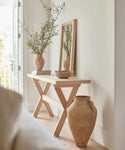 An Oak Entry Table stands against a white wall, decorated with a vase of greenery, a bowl, and a tall wooden-framed mirror; a large clay pot sits on the floor. Natural light enhances this Jenni Kayne-inspired look.