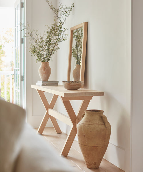 An Oak Entry Table stands against a white wall, decorated with a vase of greenery, a bowl, and a tall wooden-framed mirror; a large clay pot sits on the floor. Natural light enhances this Jenni Kayne-inspired look.