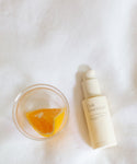 A slice of orange in a glass sits next to The Brightening Duo, creating a radiant scene on a white fabric background.