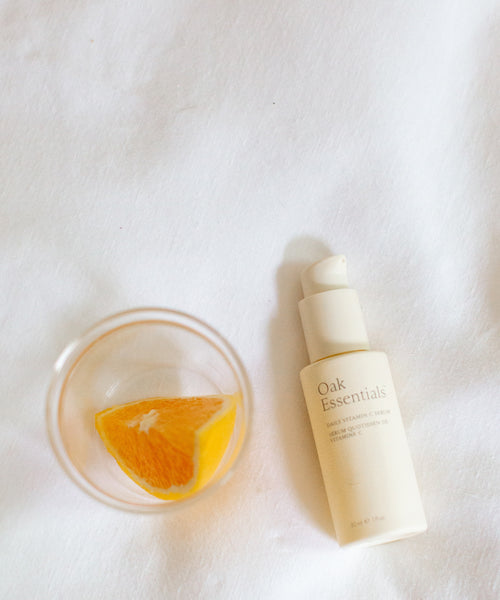 A slice of orange in a glass sits next to The Brightening Duo, creating a radiant scene on a white fabric background.
