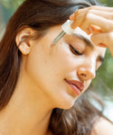 A woman with smooth skin and long brown hair uses a dropper to apply The Brightening Duo serum to her cheek near her eye, enjoying her glowing skincare ritual with a calm expression.