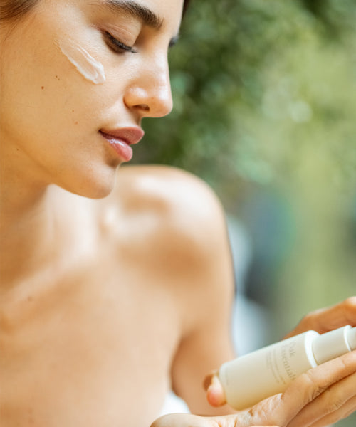 A woman with closed eyes applies cream to her cheek, holding The Brightening Duo bottle. She looks relaxed as softly blurred greenery in the background enhances the serene skincare moment.